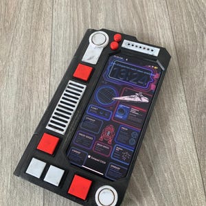 Puede incluir: Funda de teléfono negra y gris con detalles rojos y blancos, diseñada para parecerse a un panel de control. La pantalla del teléfono muestra una interfaz futurista con un gráfico de nave espacial y el texto "Datapad CX24".