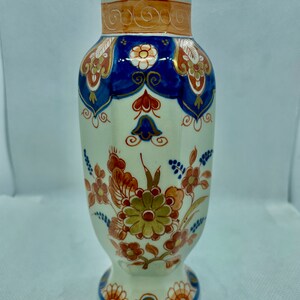 Può includere: Un vaso decorativo in ceramica con base color crema, ornato con intricati motivi floreali nei toni dell'arancione, blu e oro. Il vaso presenta una base svasata e un collo leggermente affusolato, mostrando un design tradizionale.