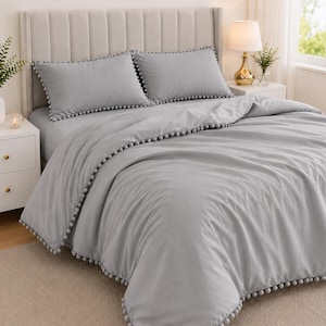 May include: A light gray bedding set featuring a duvet cover and two matching pillowcases, all adorned with a decorative pom-pom trim along the edges. The fabric has a smooth, solid-colored finish, and the pom-poms are small and spherical.
