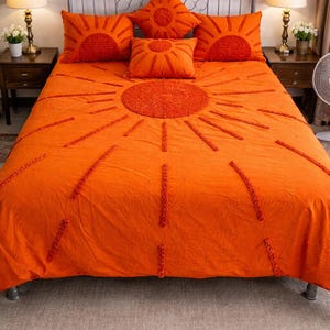 May include: Vibrant orange bedding set with a sunburst pattern. The set includes a duvet cover and several cushions, all in a bright orange colour. The sun design is created with textured, raised stitching. The bed has a metal frame.