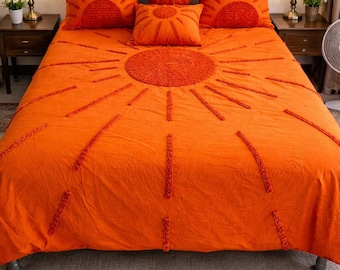 May include: Bright orange bedding set featuring a sunburst design. The set includes a comforter and several pillows, all in a vibrant orange hue. The sun design is created with textured, raised stitching. The bed is made up with a metal frame.