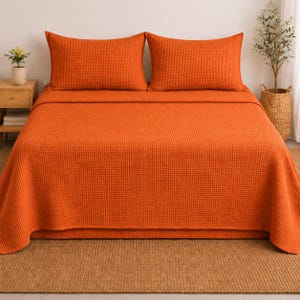 May include: An orange waffle-weave bedspread and two matching pillowcases. The bedding is neatly arranged on a bed, with a wooden nightstand and potted plant visible on the left. A woven basket with a plant is on the right.