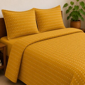 May include: A mustard yellow quilt set featuring two matching pillow shams and a bedspread. The fabric displays a consistent quilted texture with horizontal rows of small, white dashed stitching patterns across the entire surface of the bedding.