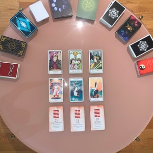 May include: A tarot card reading spread on a pink table with a variety of cards, including the Justice card, the Two of Cups card, the Two of Wands card, and the Eight of Swords card. The spread also includes three cards from the I Ching, which are numbered 21, 11, and 53.