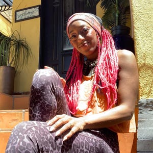 May include: A person with bright pink braided hair wearing a patterned headscarf, an orange sleeveless top, and dark gray leggings with a black leopard print pattern. The person is adorned with a silver necklace and a ring on their finger.