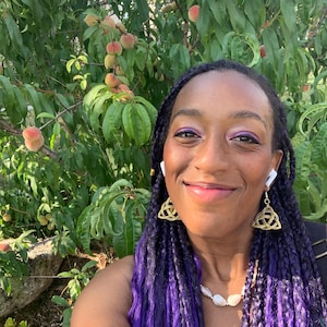 May include: A woman with purple braids and gold Celtic knot earrings smiles in front of a peach tree. She wears a white pearl necklace and white earbuds. Ripe peaches are visible on the tree in the background.