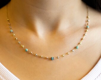 May include: A gold chain necklace with small turquoise and black beads. The necklace is worn by a model.