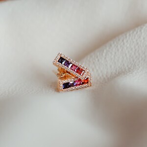 May include: Gold bar earring featuring a row of colourful square gemstones. The gemstones are set in a gold frame with a row of smaller clear gemstones around the edge.