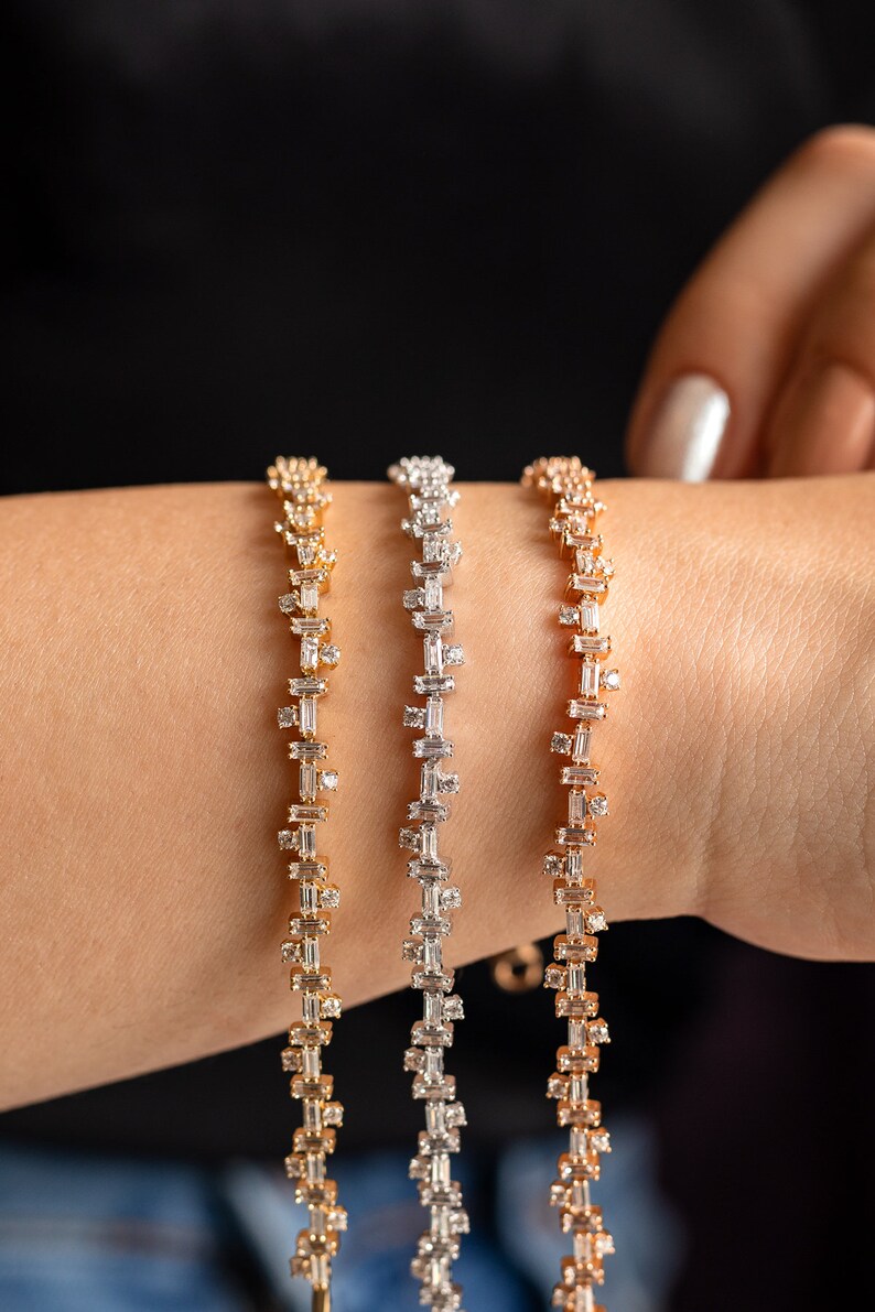 May include: Three gold, silver, and rose gold bracelets with baguette-shaped diamonds. The bracelets are stacked on a person's wrist.