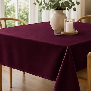 May include: A square, deep burgundy tablecloth with a smooth, solid-colored fabric texture draped over a table. The tablecloth features clean, finished edges and hangs evenly on all sides, providing a rich, dark color contrast to the light wood furniture.