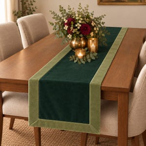 Puede incluir: Un camino de mesa rectangular de terciopelo con un panel central de color verde bosque profundo bordeado por un ribete de color verde salvia más claro. El camino está extendido sobre una mesa de comedor de madera, con textura suave.