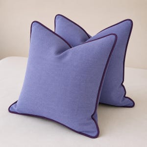 Peut inclure: Deux coussins carrés en tissu bleu pervenche uni. Chaque coussin présente un passepoil violet foncé distinct le long des quatre bords, offrant une bordure nette et définie à la surface texturée des housses de coussin.