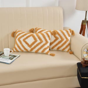 May include: Two rectangular throw pillows with white fabric and a geometric pattern in a golden-yellow color. Each pillow has tassels at the corners. The pillows are on a beige sofa.