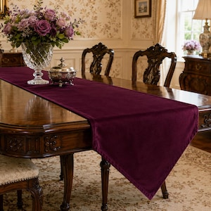 May include: A deep plum-colored velvet table runner with a rectangular shape and pointed ends, draped lengthwise across the center of a polished wooden dining table, providing a soft, textured surface for decorative items like a glass vase.