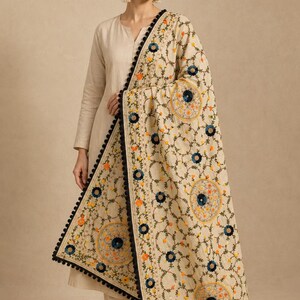 Könnte beinhalten: Ein beigefarbener Dupatta mit aufwendiger Blumenstickerei in Dunkelblau, Orange und Gelb. Der rechteckige Stoff ist mit einer schwarzen Bommelborte eingefasst und zeigt durchgehend kreisförmige Muster auf dem hellen Untergrund.