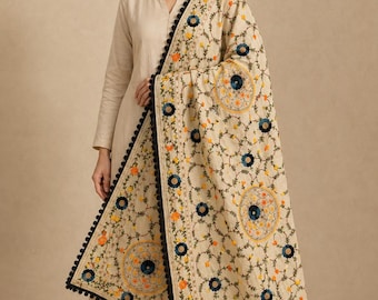 May include: A beige dupatta featuring intricate floral embroidery in shades of dark blue, orange, and yellow. The rectangular fabric is bordered with a black pom-pom trim and displays circular patterns throughout the design on a light base.