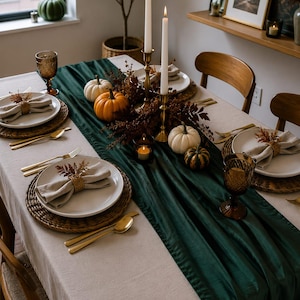 May include: A dining table set with a beige tablecloth and a dark green velvet table runner. The setting includes white plates on woven chargers, gold-toned cutlery, amber-coloured glasses, linen napkins with dried foliage, and candles.