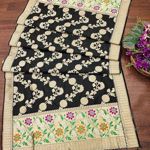 May include: A black rectangular textile featuring a central pattern of repeating cream-coloured floral motifs. The ends are bordered by wide bands of cream fabric decorated with green vines, orange flowers, and magenta blossoms, finished with black tassels.