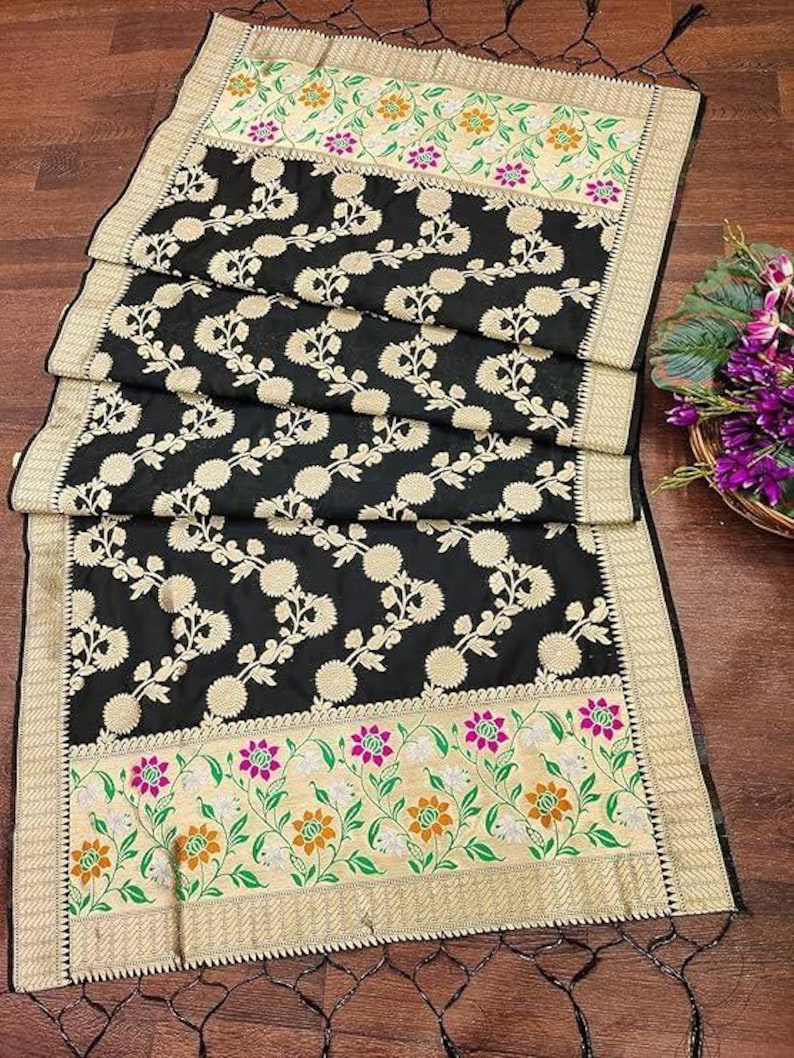 May include: A black rectangular textile featuring a central pattern of repeating cream-coloured floral motifs. The ends are bordered by wide bands of cream fabric decorated with green vines, orange flowers, and magenta blossoms, finished with black tassels.
