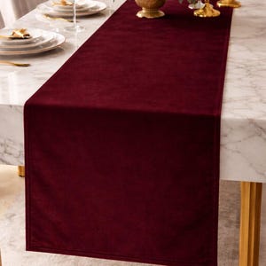 May include: A burgundy velvet table runner displayed on a marble table. The runner is rectangular and covers the center of the table. The table is set with plates, glasses, and a floral centerpiece.