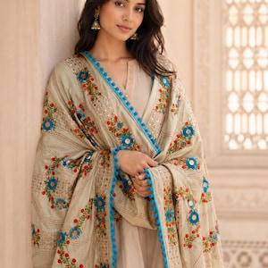 May include: A beige embroidered shawl with blue, red, and green floral patterns. The shawl has a blue pom-pom trim and mirror accents. The woman is wearing a beige outfit and earrings.