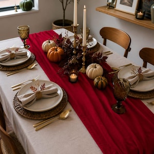 Pode incluir: Uma mesa de jantar com toalha bege e um caminho de mesa vermelho profundo. O conjunto inclui pratos brancos sobre bases trançadas, guardanapos bege com folhas, talheres dourados, copos âmbar, castiçais de latão e pequenas abóboras.