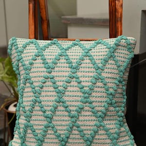 May include: A square throw pillow with a geometric pattern in shades of teal and white. The pillow is made of a textured fabric with raised bumps.