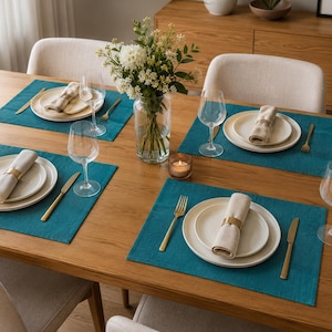 Può includere: Un tavolo da pranzo apparecchiato per quattro con tovagliette rettangolari color ottanio, piatti in ceramica color crema, tovaglioli di lino beige con anelli dorati, posate dorate, calici da vino e un vaso di vetro con fiori bianchi.