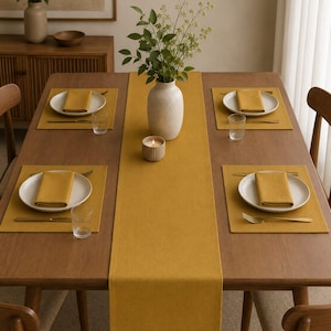 Op de afbeelding: Een rechthoekige houten eettafel met een mosterdgele stoffen tafelloper en vier bijpassende placemats. Elke plaatsinstelling bevat een wit keramisch bord, een gevouwen mosterdgele servet, een glazen beker en een goudkleurige vork.