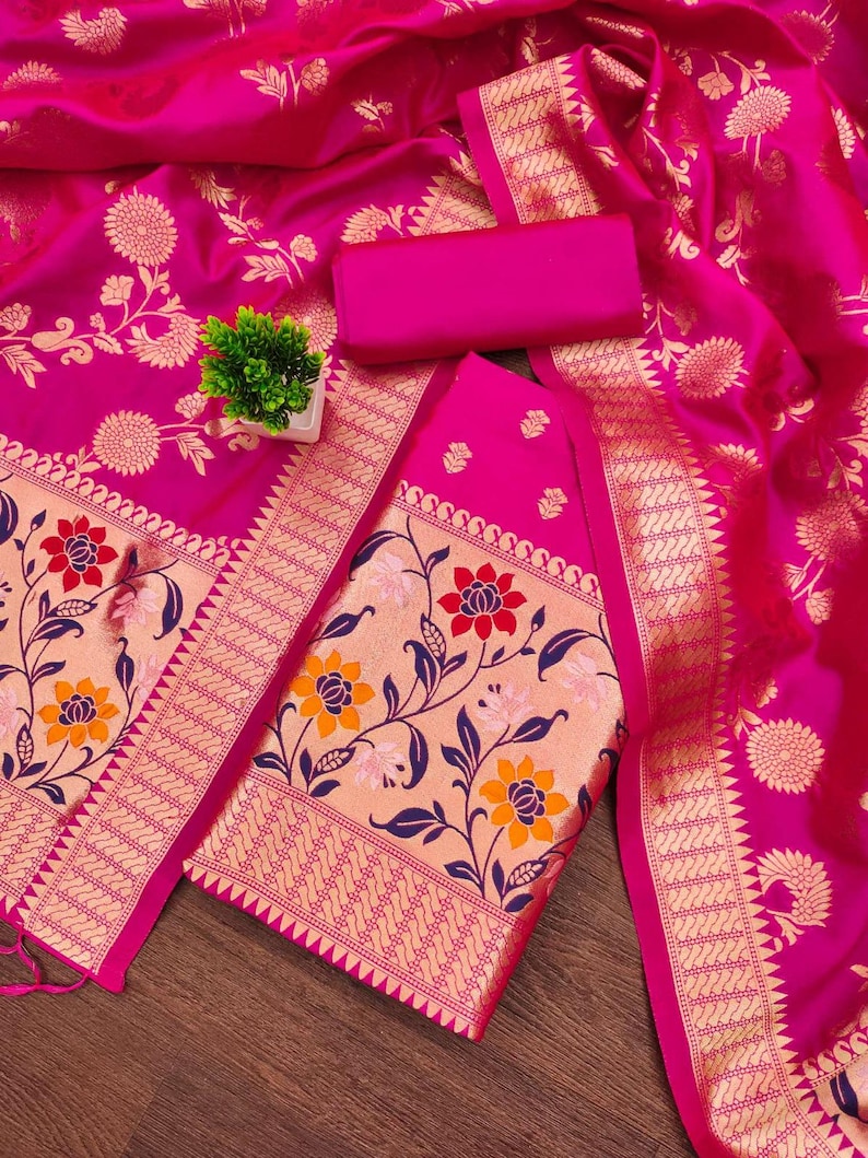 May include: A vibrant fuchsia fabric featuring intricate gold floral patterns and a wide border with detailed botanical motifs in red, orange, and dark blue. The textile includes a matching solid fuchsia rectangular piece and a small green plant.