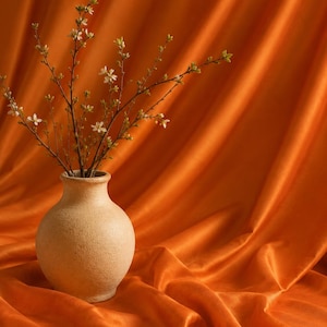 以下が含まれることがあります： 細い首を持つベージュ色の質感のある陶器製の花瓶に、小さな緑の葉と繊細な白い花がついた数本の細い茶色の枝が生けられており、柔らかなひだのある鮮やかなオレンジ色の布を背景に配置されています。