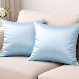May include: Two pale blue satin cushions are placed on a beige sofa. The cushions have a smooth, shiny texture and are square. A vase of white flowers sits on a side table in the background.