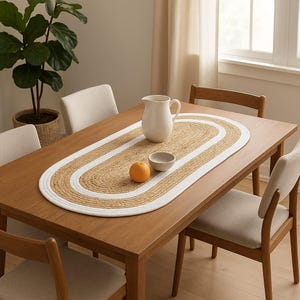 May include: An oval table runner made of braided natural jute fiber with a thick white border. The runner is placed on a wooden dining table and features a textured, woven surface with a distinct contrast between the tan and white materials.
