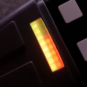 May include: Close-up of a black electronic device with a glowing yellow and red bar display. The device has several square buttons in white and red. The overall design is sleek and modern.