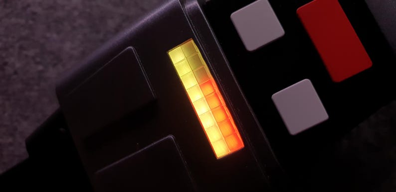 May include: Close-up of a black electronic device with a glowing yellow and red bar display. The device has several square buttons in white and red. The overall design is sleek and modern.