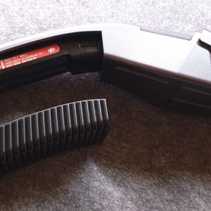 May include: A futuristic-looking, silver and black device with a curved handle and a rectangular body. A separate, black, ridged component sits beside it. The device has a red sticker with white text. The overall design suggests a prop or gadget.