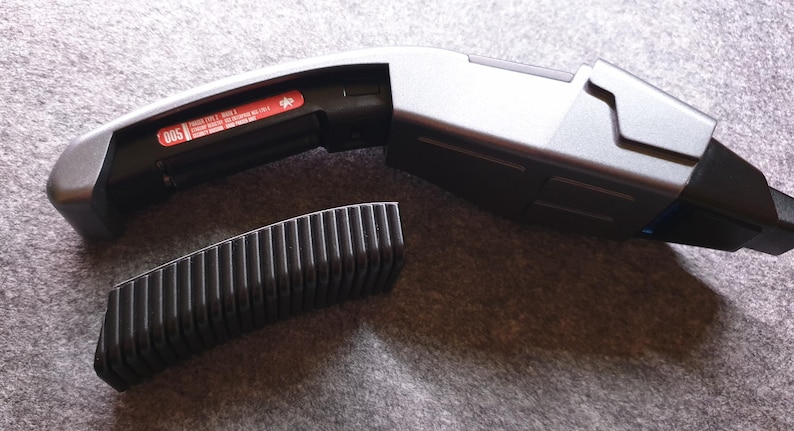 May include: A futuristic-looking, silver and black device with a curved handle and a rectangular body. A separate, black, ridged component sits beside it. The device has a red sticker with white text. The overall design suggests a prop or gadget.