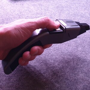 May include: A gray and black prop phaser, a fictional handheld weapon, is held in a hand. The phaser has a rectangular body with a curved handle. The person's thumb is pressing a button on the top of the phaser.