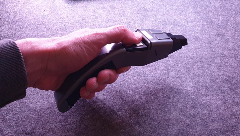 May include: A gray and black prop phaser, a fictional handheld weapon, is held in a hand. The phaser has a rectangular body with a curved handle. The person's thumb is pressing a button on the top of the phaser.