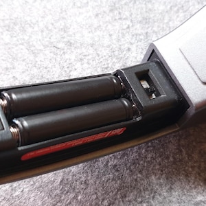 May include: Close-up of a silver and black electronic device with a battery compartment open, revealing two black AA batteries. The device has a sleek, modern design with a rectangular shape and a matte finish.