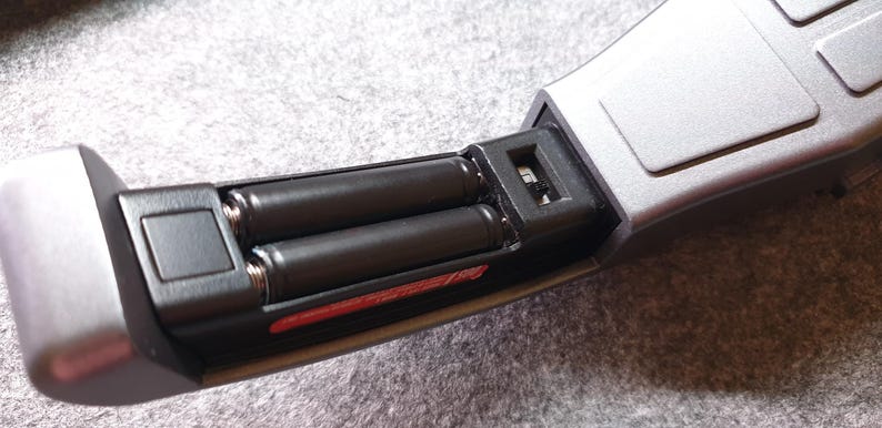 May include: Close-up of a silver and black electronic device with a battery compartment open, revealing two black AA batteries. The device has a sleek, modern design with a rectangular shape and a matte finish.