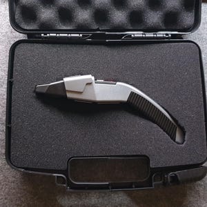 May include: A silver and black device with a curved handle rests inside an open black case with foam padding. The device has a futuristic design, with a silver top and a black, textured grip. The case is rectangular with a hinged lid.