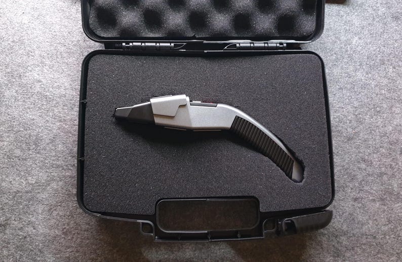 May include: A silver and black device with a curved handle rests inside an open black case with foam padding. The device has a futuristic design, with a silver top and a black, textured grip. The case is rectangular with a hinged lid.