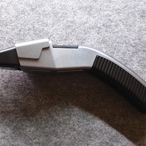 May include: A silver and black prop device with a curved handle and a rectangular head. The handle has a ridged texture for grip. The device is likely a prop from a science fiction series.