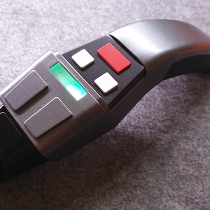 May include: A gray and black prop device with a curved handle and a rectangular head. The head features a green illuminated display, two gray rectangles, two white squares, and a red rectangle. The device is likely a futuristic prop.