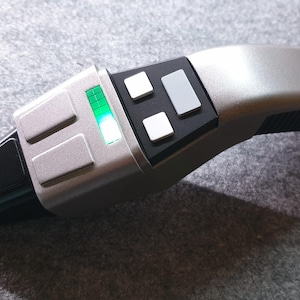 May include: A handheld electronic device with a curved silver and black casing. The device features a series of rectangular buttons, a black grip section, and a small rectangular display screen that emits a bright green light when activated.