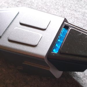 May include: A close-up of a silver and black USB-C adapter. The adapter has a rectangular shape with a series of raised rectangular details on the top. A blue light is visible on the end of the adapter.