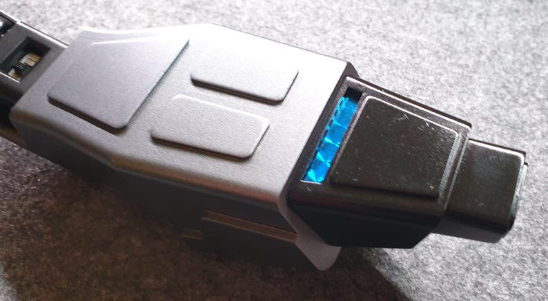May include: A close-up of a silver and black USB-C adapter. The adapter has a rectangular shape with a series of raised rectangular details on the top. A blue light is visible on the end of the adapter.