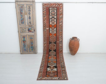 May include: A long, narrow runner rug with a traditional geometric pattern in shades of red, orange, black, and white. The rug has a fringe on both ends.