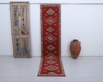 May include: A long, rectangular red rug with geometric patterns in beige and brown. The rug is displayed vertically against a white wall. An antique wooden door with colorful painted designs and a large brown vase are also in the image.